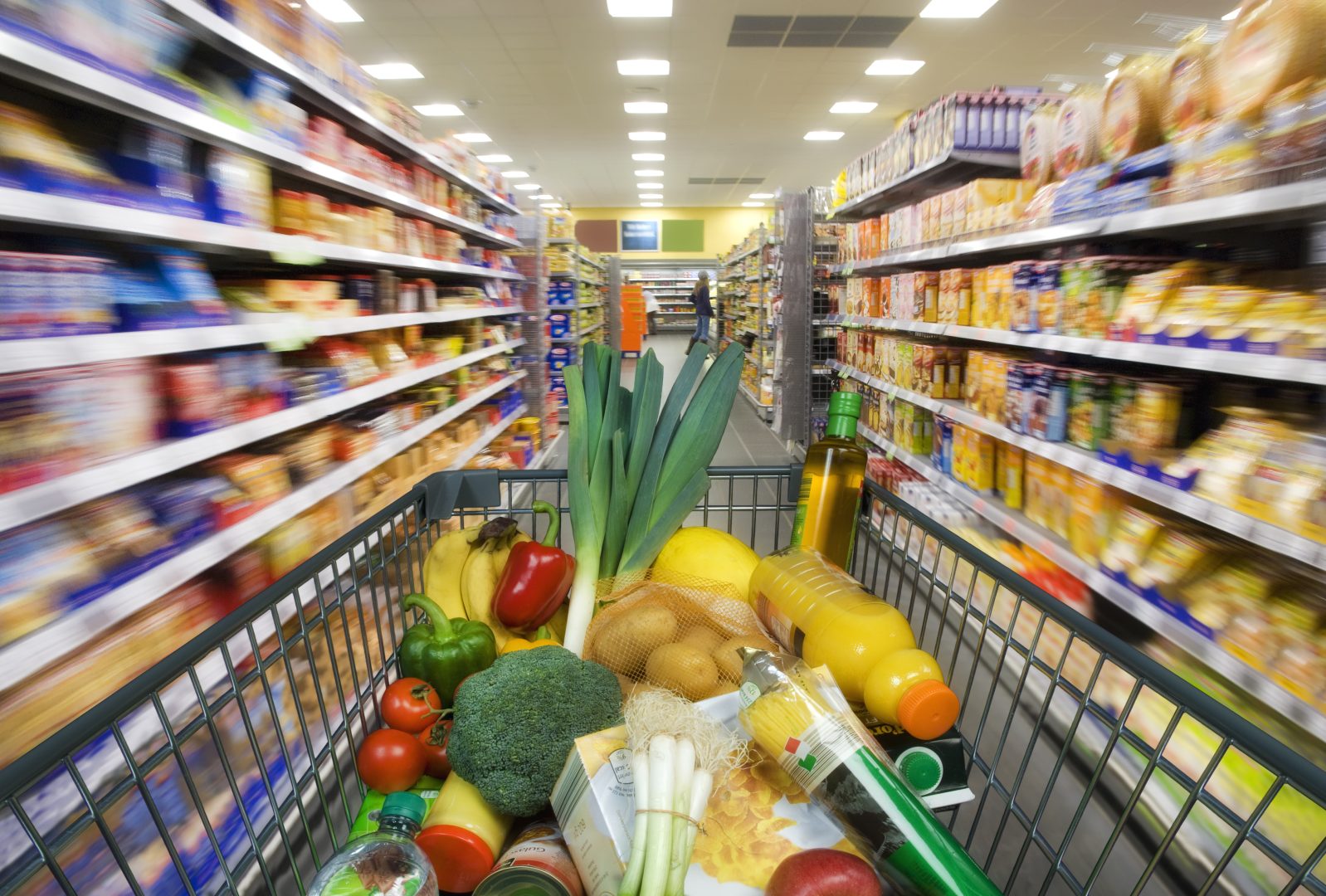 Perspective from a grocery cart showing fresh produce in the basket with blurred store shelves in the background, suggesting shopping in a supermarket.